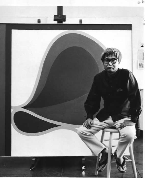 Matsumi Kanemitsu seated on a stool in front of one of his paintings.