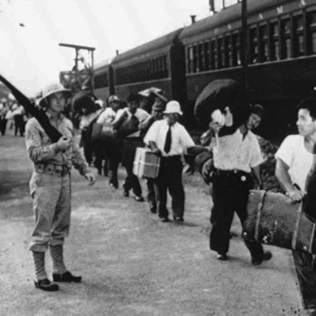 Japanese Latin Americans hold suitcases in a line while a soldier holds a gun.