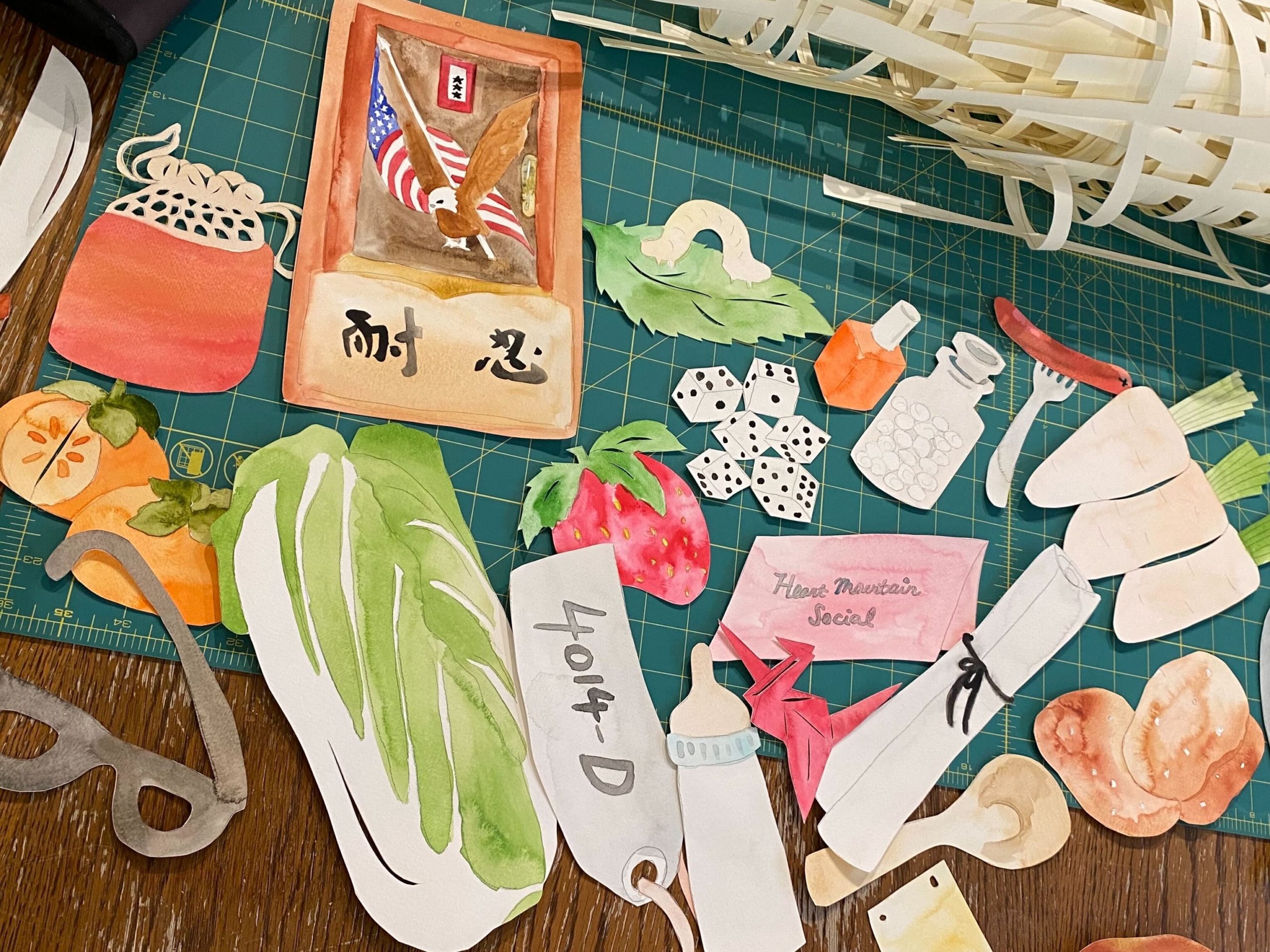 Objects cut from paper and painted in watercolor, laid out on a table before being added to the memory net. Some of the objects include a head of napa cabbage, an evacuation tag, origami crane, wooden rice paddle, baby bottle, and a silkworm on a mulberry leaf.