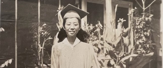 Sarah Yomogi Okada standing in front of a barrack in Jerome concentration camp wearing a graduation cap and gown.