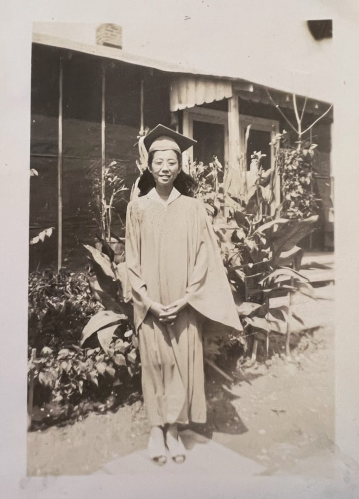 Sarah Yomogi Sato standing in front of a barrack in Jerome concentration camp wearing a graduation cap and gown.