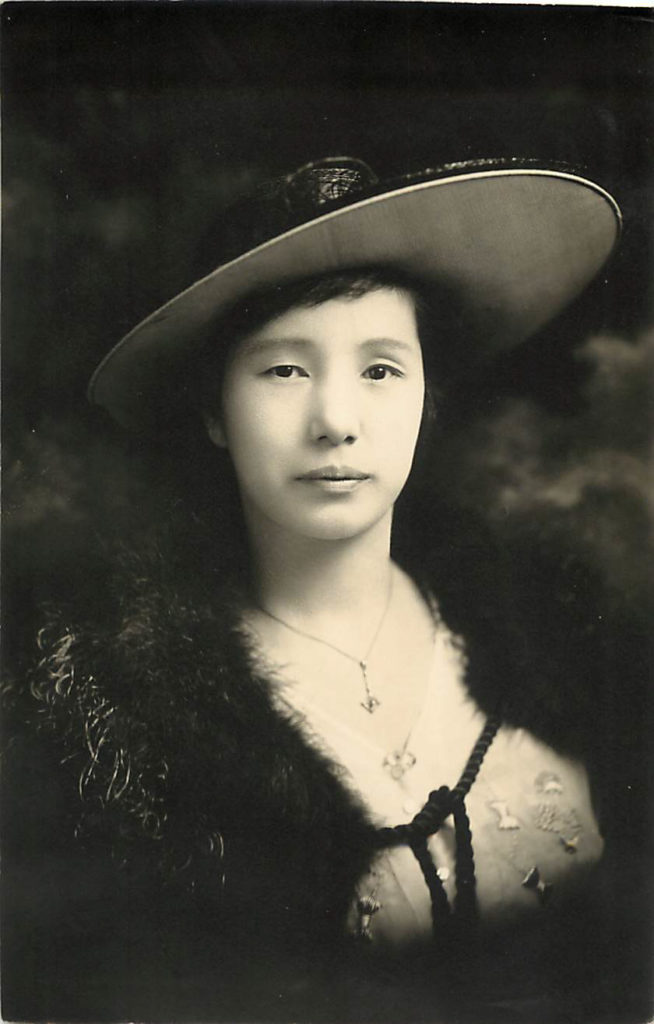 A young Aya Hori Masuoka circa 1910. She is wearing western clothes and a hat and pictured from the chest up with a slight smile.