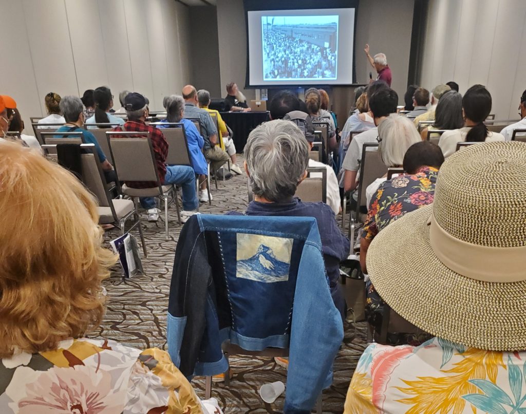Heart Mountain survivor Sam Mihara giving a presentation to a room of people. One participant in the foreground has a denim jacket with a sketch of Heart Mountain and sashiko stitching draped over the back of their chair.