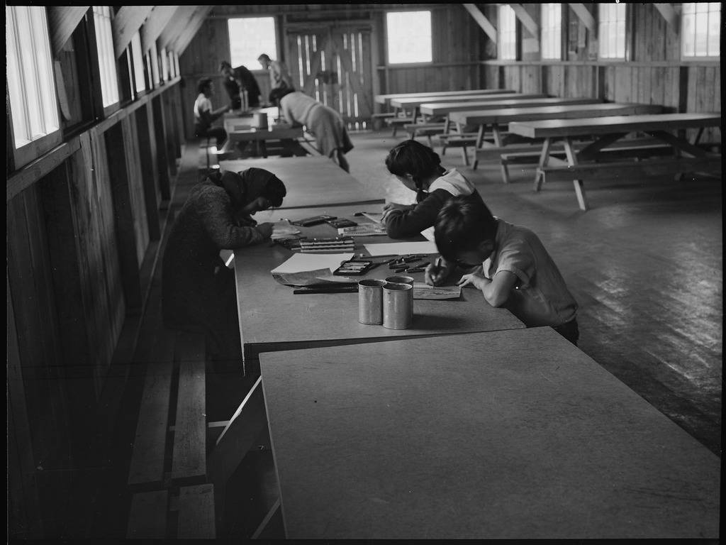 Three Japanese American children sitting around a table working on artwork after a class at the Tanforan Art School.