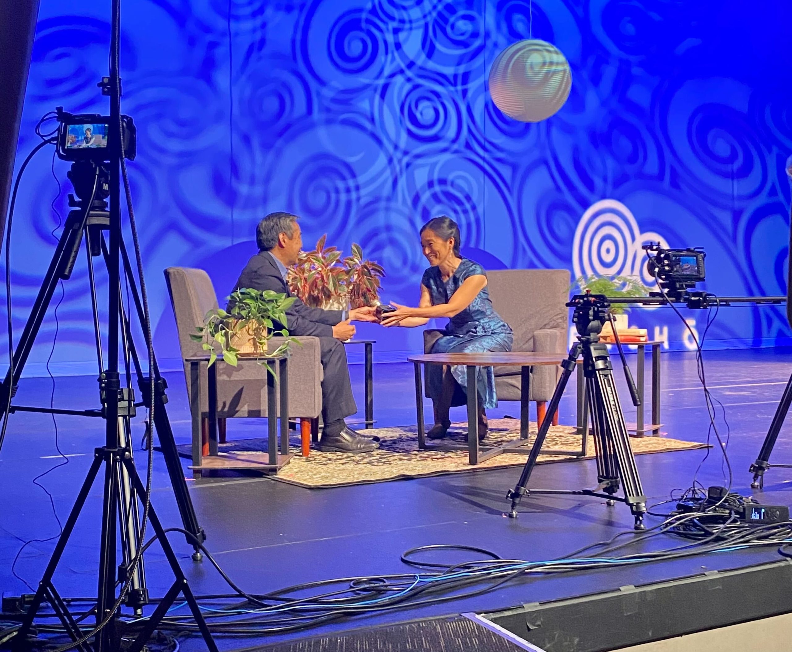 Tom Ikeda and Naomi Ostwald Kawamura in conversation seated on stage during filming of the Densho Virtual Gala. Ikeda is handing off a baseball trophy to Naomi that was gifted to him by a Nisei community leader when he first started at Densho.