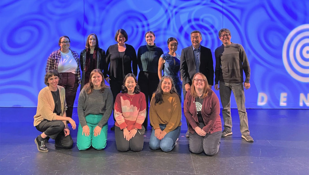Densho staff pose for a group photo after the gala. They are on a stage with a blue background and the Densho logo, standing and kneeling in two rows.