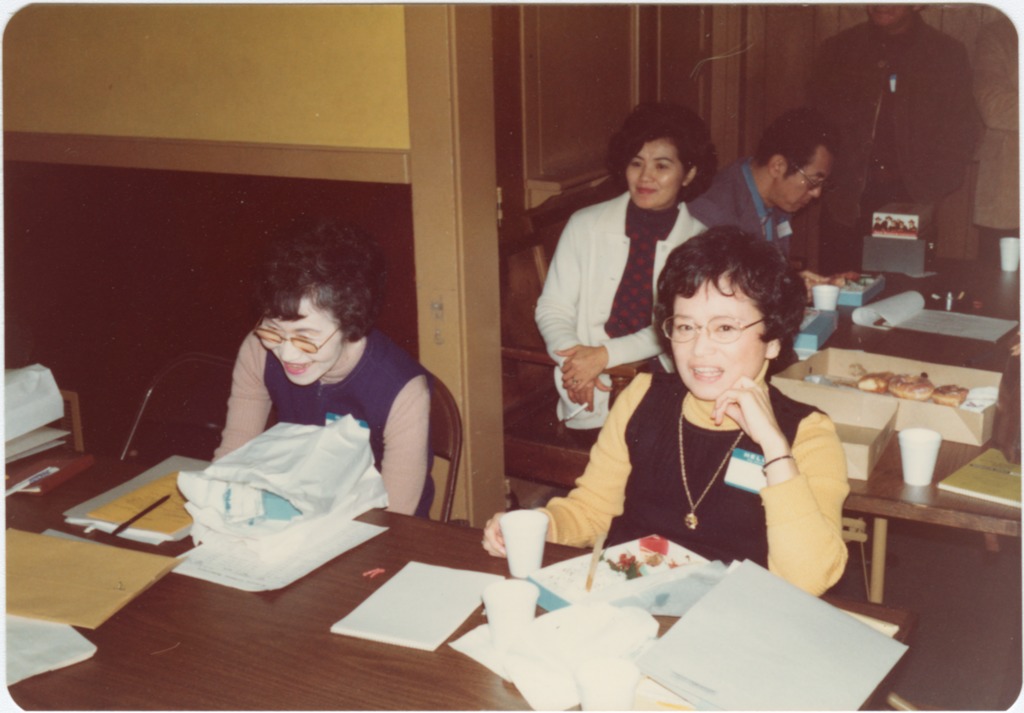 Cherry Kinoshita at a JACL meeting in 1976. She is seated next to another woman at a table with notepads and styrofoam cups. Behind them are two more people working at another table.