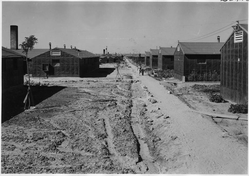 A mother and child walking down a dirt path between barracks at Minidoka concentration camp.