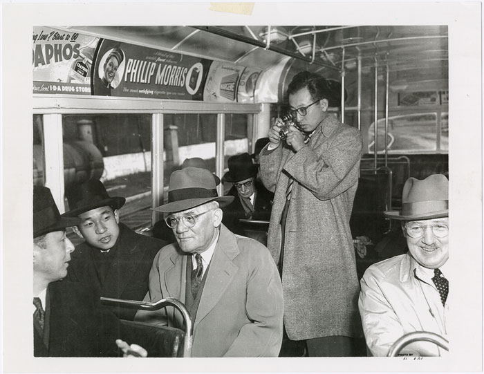 Hikaru Iwasaki standing in the middle of a bus taking a photo of Prince Akihiro, who is seated talking to three men, in 1953.