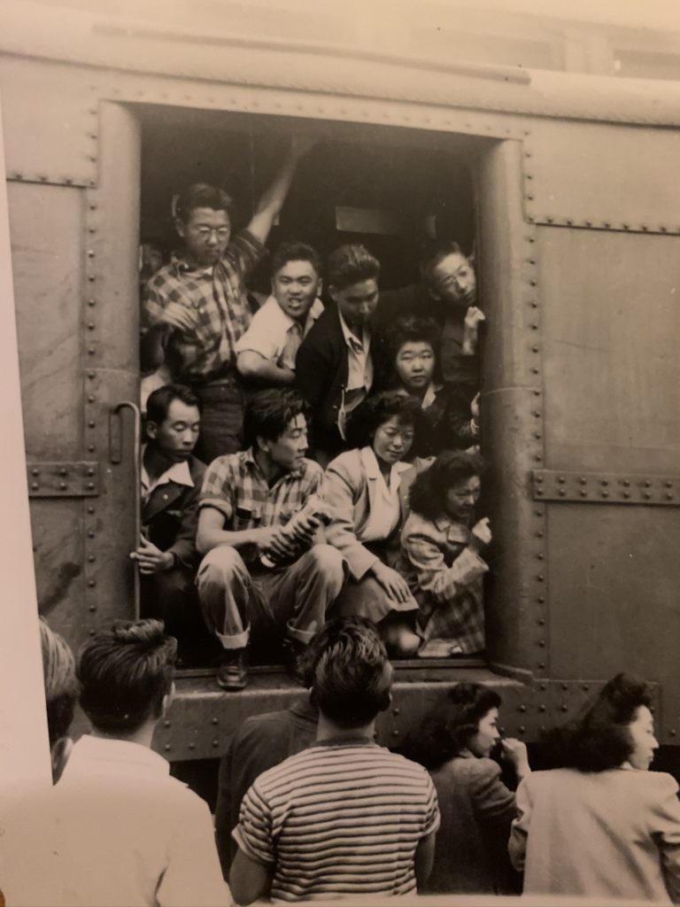 Nine Japanese American young adults crowded into the doorway of a train car, saying goodbye to friends standing on the ground in front of the train.