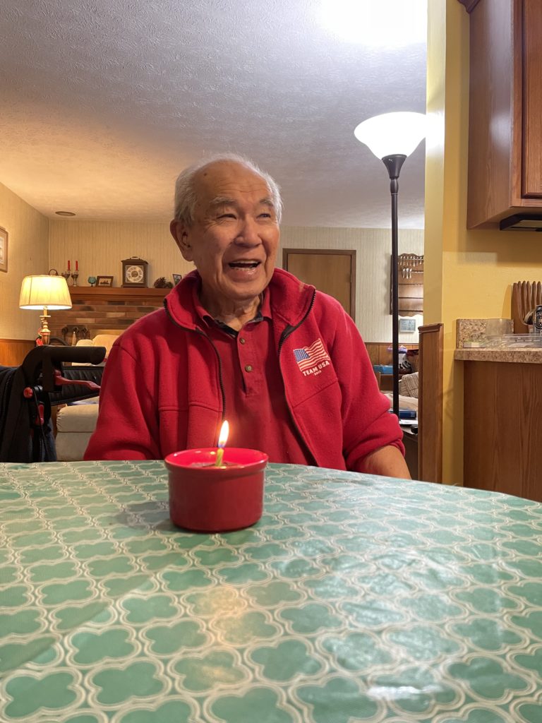 Kayoshi Masuoka celebrating his 95th birthday. He has a big smile and is seated at a kitchen table next to a cupcake with a lit candle.