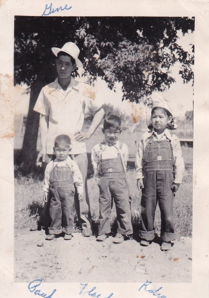 A Japanese American man standing outdoors with his three young children. The children appear to be wearing matching shirts and overalls.