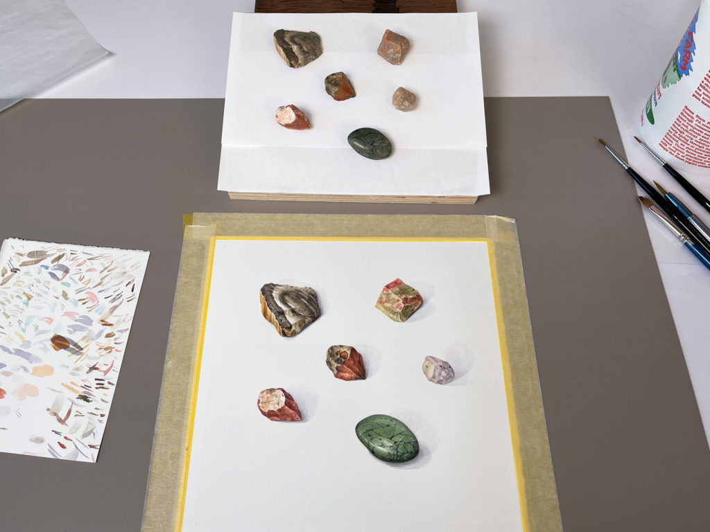 A watercolor in progress from Alison Moritsugu's "Moons and Internment Stones" exhibition. The actual rocks being painted are shown next to the painting.