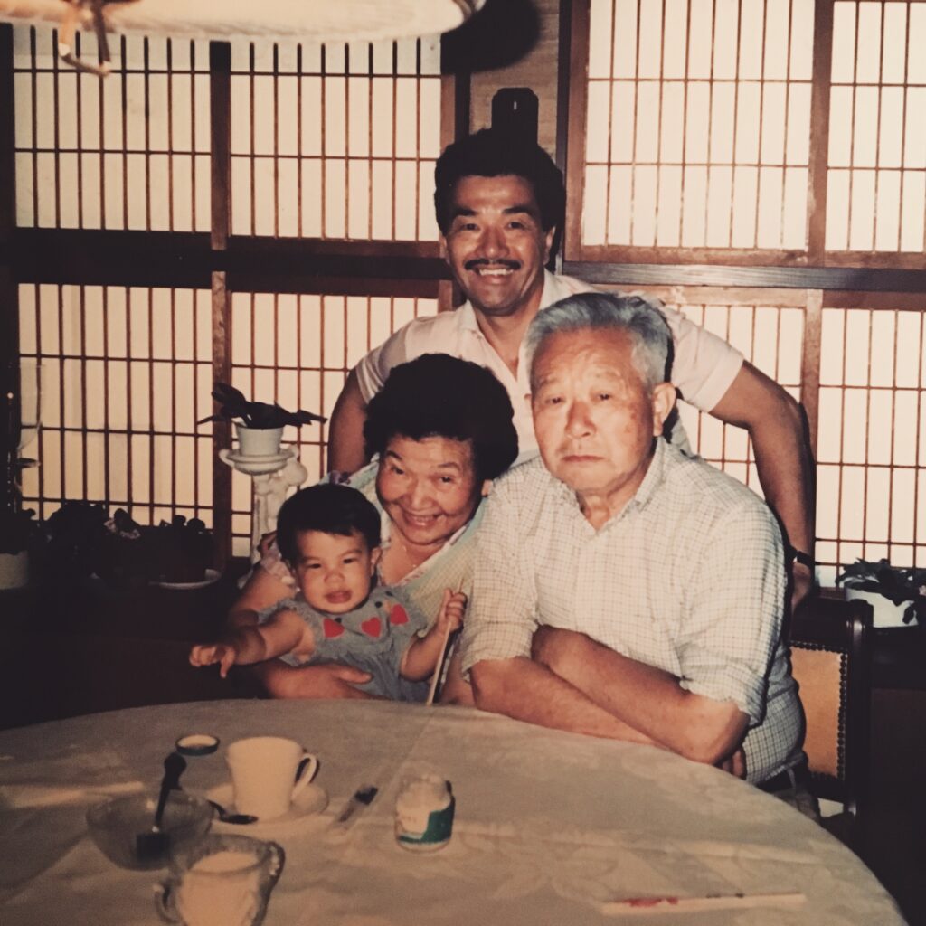 Diana Tsuchida as a baby with her father and grandparents. Her grandmother is holding Diana in her lap, sitting at a table next to her grandfather, while her father stands behind them.
