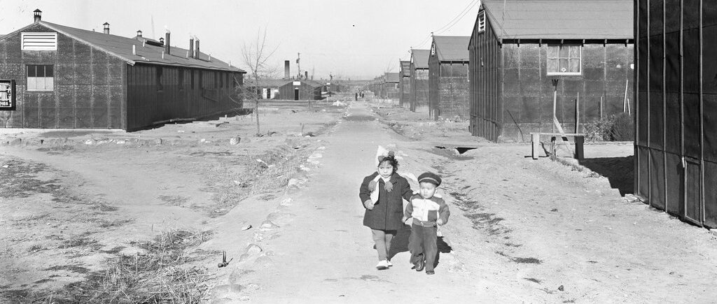 Two small children walking down a dirt path between barracks at Minidoka.
