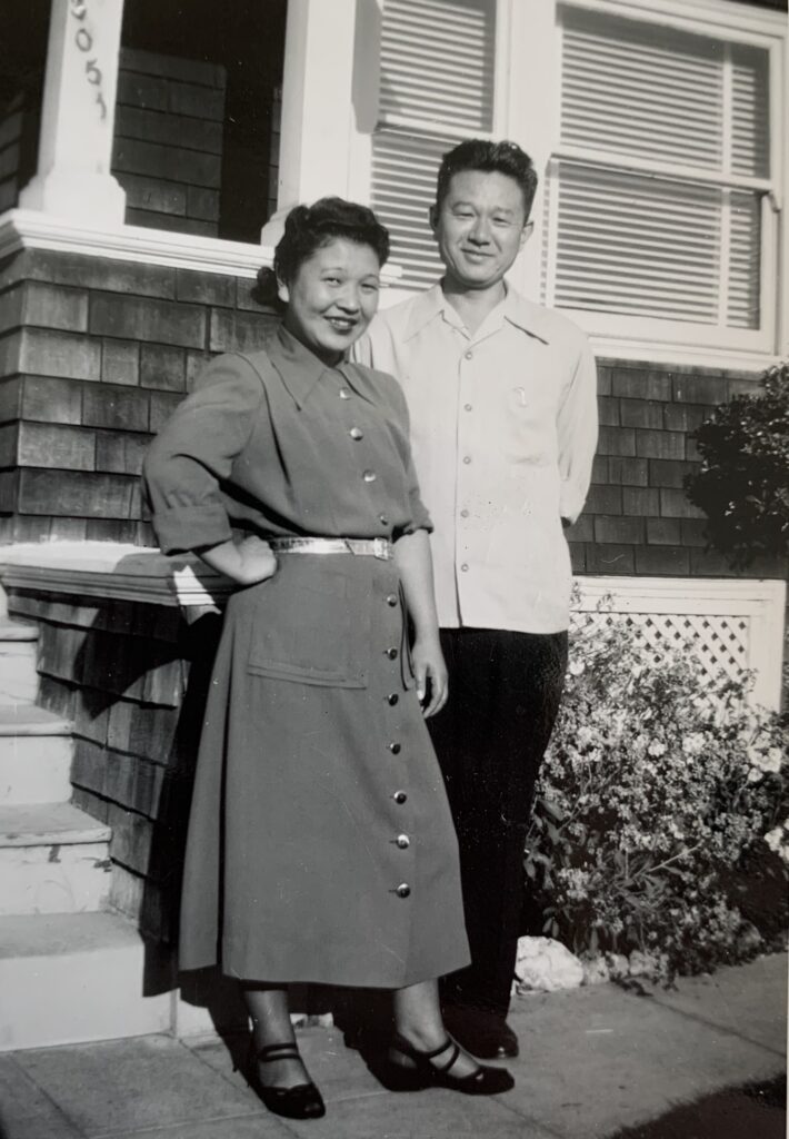 Tamotsu and Itsuye Tsuchida standing in front of a home in Berkeley, California. They are standing close together, smiling at the camera.