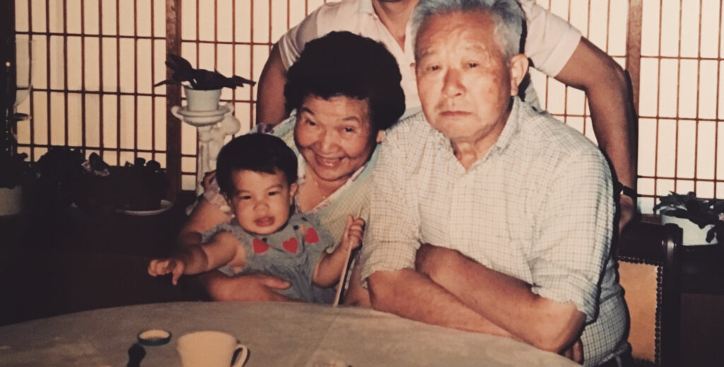 Diana Tsuchida as a baby with her father and grandparents. Her grandmother is holding Diana in her lap, sitting at a table next to her grandfather, while her father stands behind them.