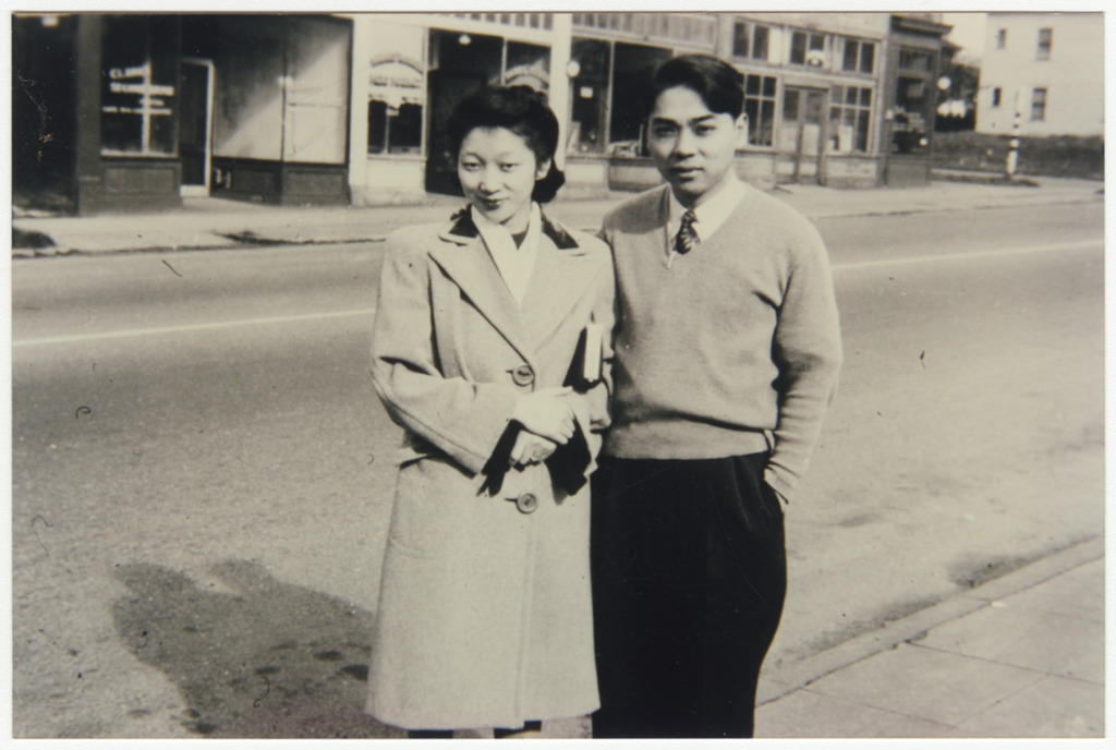 Tama and George Tokuda in Seattle circa 1946. They are standing arm in arm on a sidewalk next to an empty street.