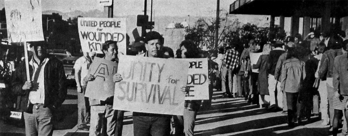 People marching with signs at the Wounded Knee occupation. Signs read "Unity for Survival" and "United People for Wounded Knee."