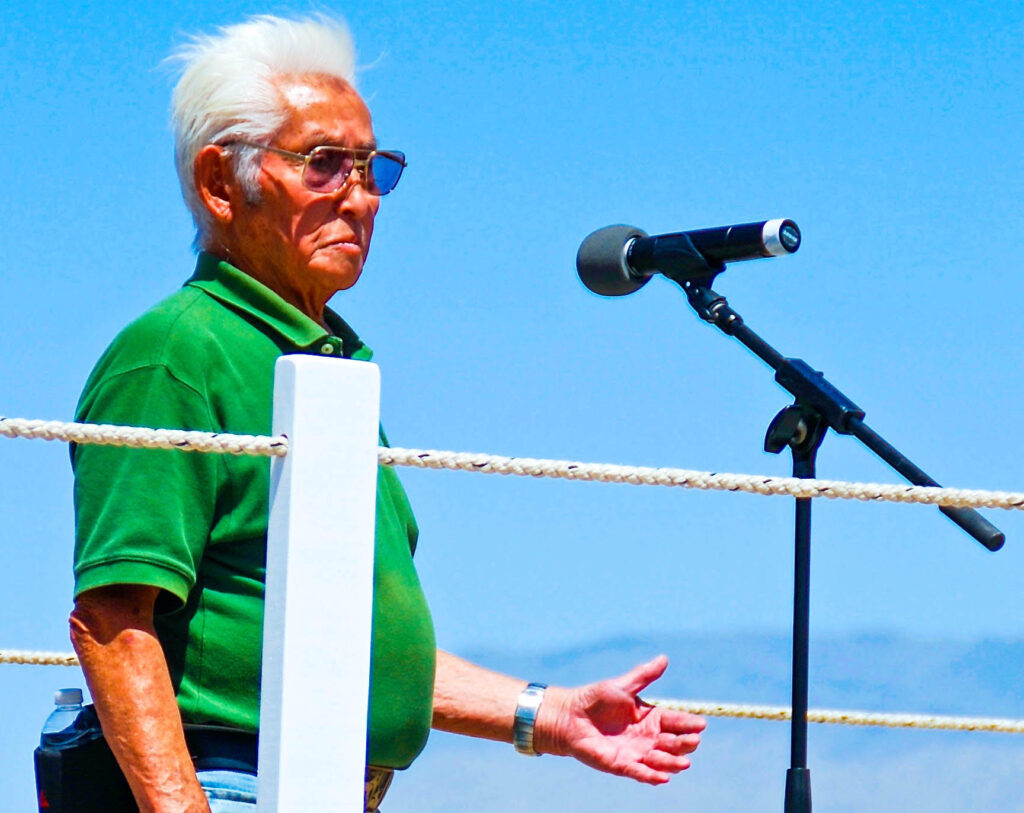 Hank Umemoto speaking at the Manzanar Pilgrimage in 2013. He is standing behind a microphone, wearing a green polo shirt and tinted aviator glasses.