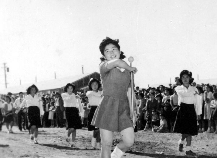 A Japanese American teen performing a flag salute during an Independence Day parade at Tule Lake. She has a big smile and is holding a baton and leading the parade.
