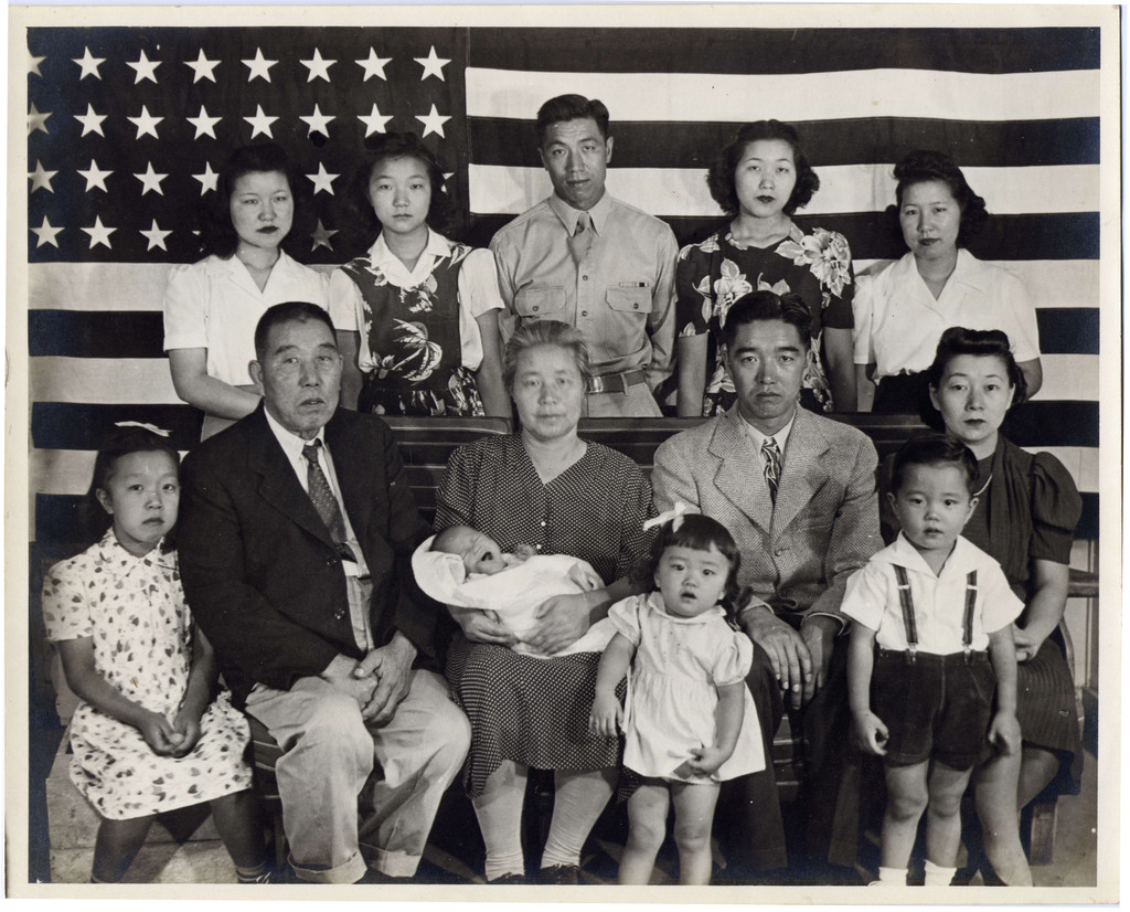 A Japanese American family posing in front of an American flag inside a WWII concentration camp. There are thirteen people ranging in age from a baby to two grandparents.