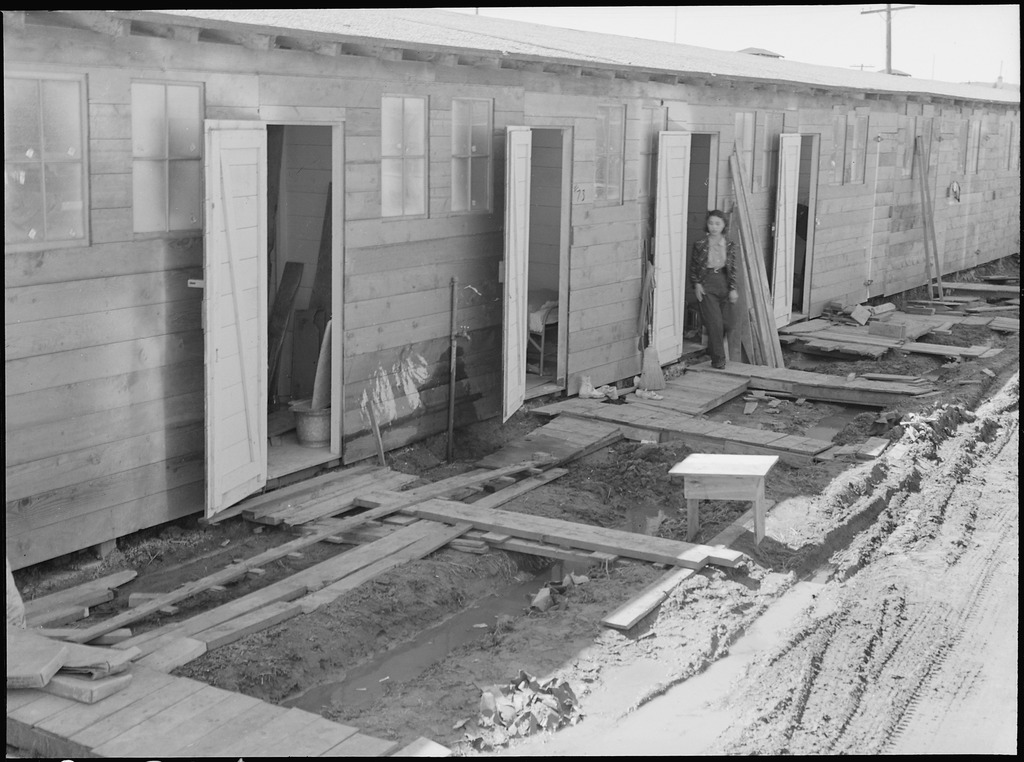 A Japanese American walking out of a barrack at the Tanforan Assembly Center. They are walking on scraps of wood laid the ground to avoid very muddy conditions.