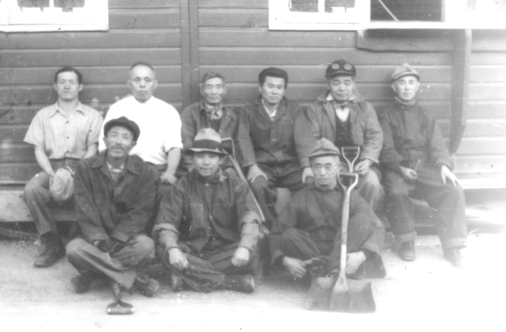 Nine Japanese American laborers who worked on a coal crew at Heart Mountain. They are sitting outside a barrack in two rows, some holding shovels.