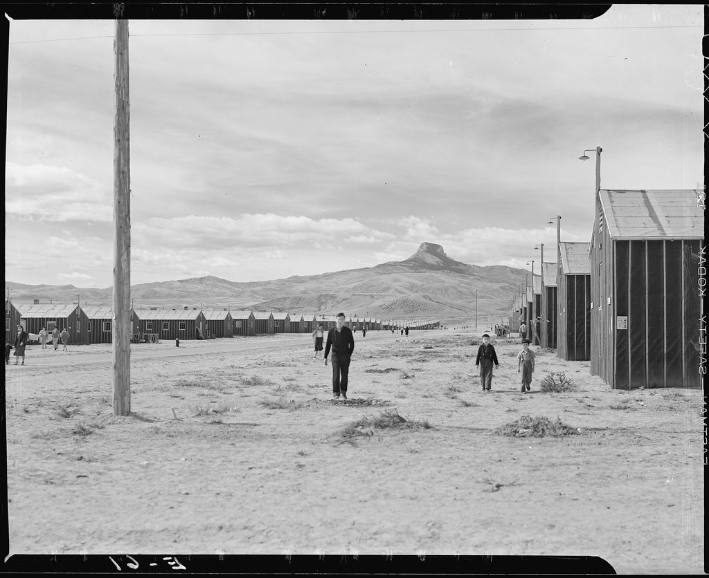 Japanese Americans walking in a street between barracks at Heart Mountain concentration camp. Heart Mountain is visible in the distance.