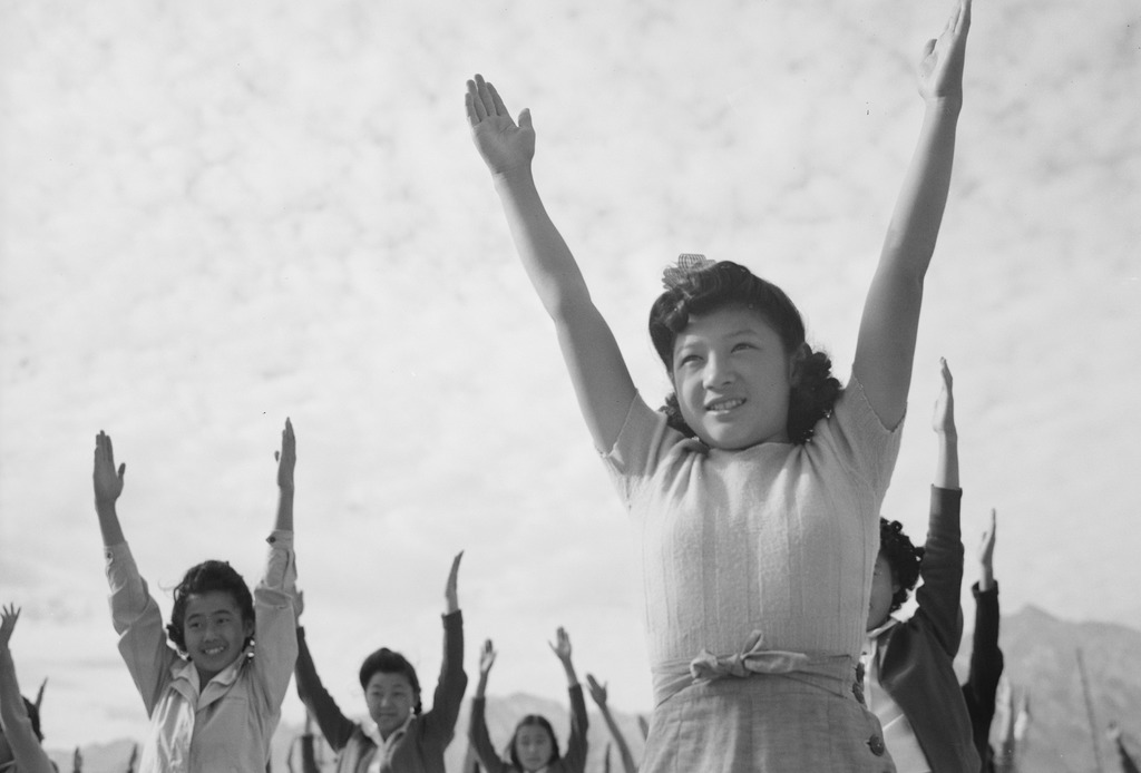 Japanese Americans participating in an exercise class at Manzanar concentration camp. They are standing in rows with their arms raised to the sky and smiling.
