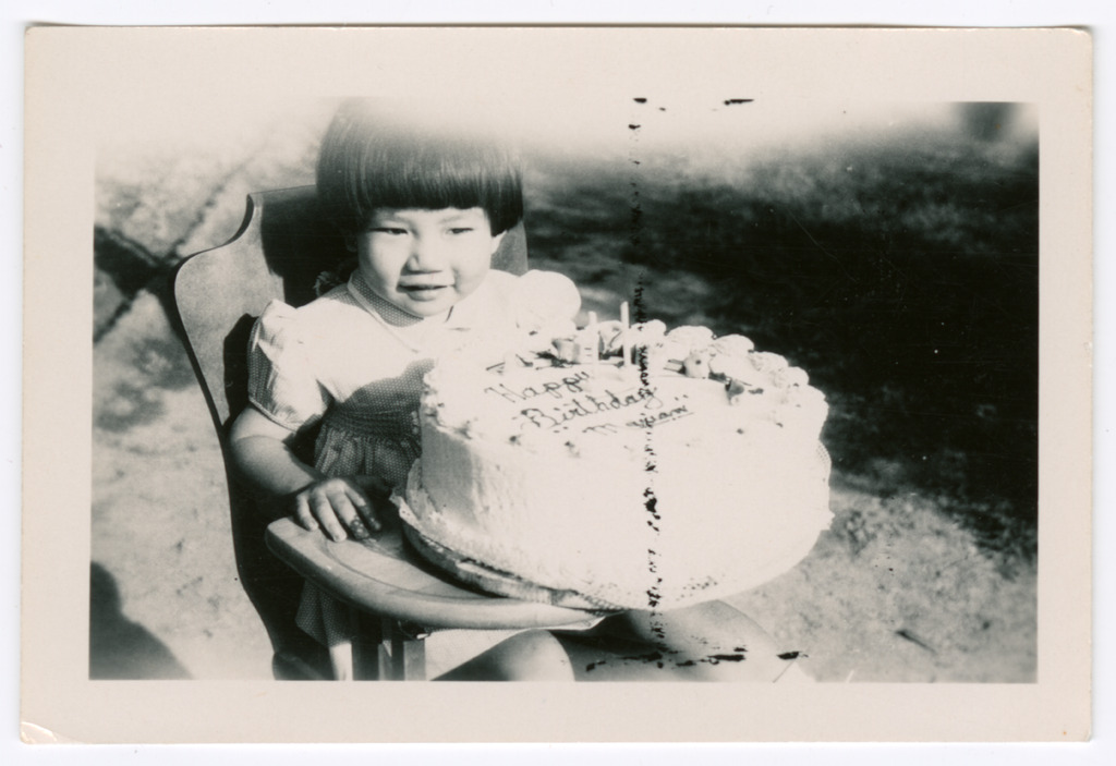 Black and white photograph of a toddler in a high chair with a large birthday cake with two candles that reads "Happy Birthday Marian"