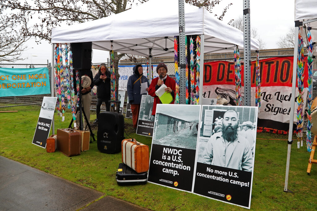 Activist Stan Shikuma speaking at a rally outside the Northwest Detention Center. In front of him are posters with photos of Japanese American incarceration camps that say 