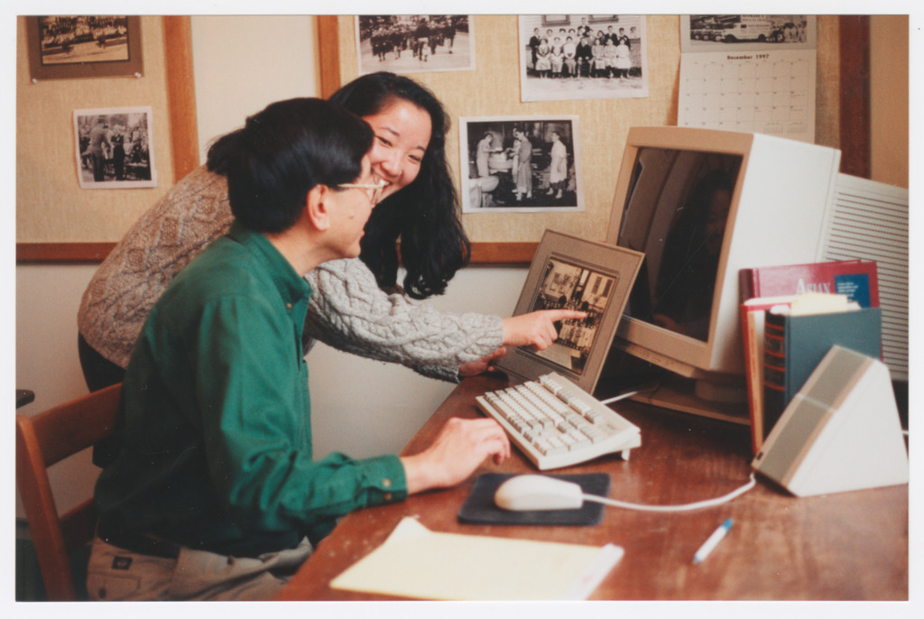 Two Densho staff members at a computer workstation looking at a printed photograph
