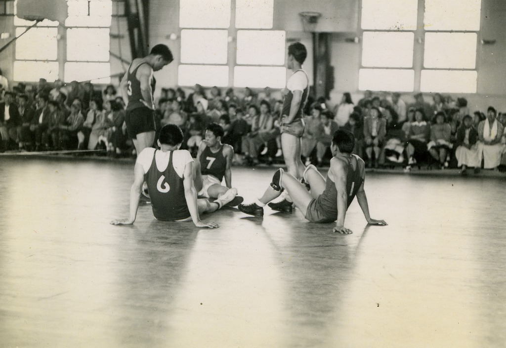 Five Japanese American players on a basketball court during a game, while a large crowd watches from the sidelines.