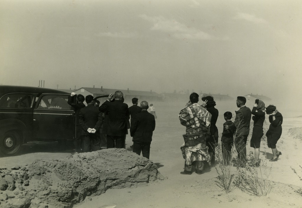 Memorial service for an Amache serviceman killed in action. Barracks are visible in the background and attendees are attempting to cover their faces as the wind blows sand over them.