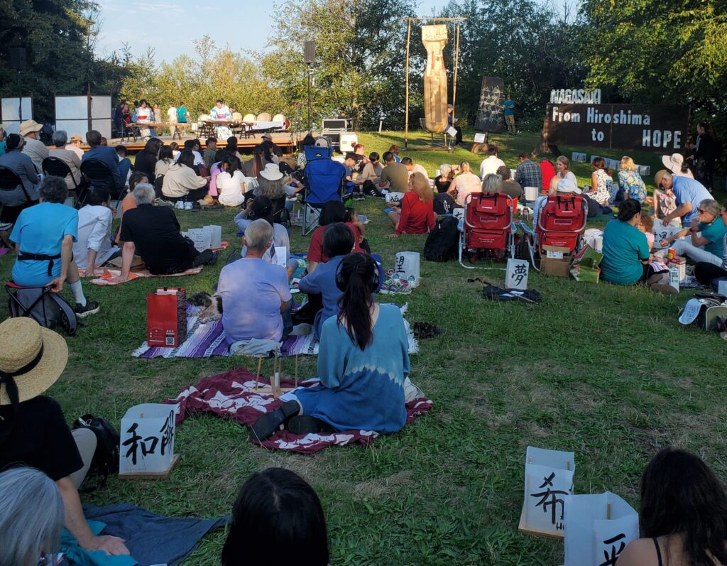 People seated on the grass in front of a small stage to watch a koto performance during the From Hiroshima to Hope ceremony. A large wooden sign off to the side says "From Hiroshima/Nagasaki to Hope."