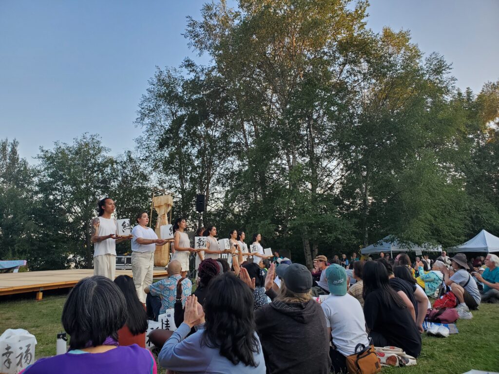 Eight dancers perform a piece at From Hiroshima to Hope. They are wearing white and standing in front of the crowd holding up paper lanterns with outstretched hands.