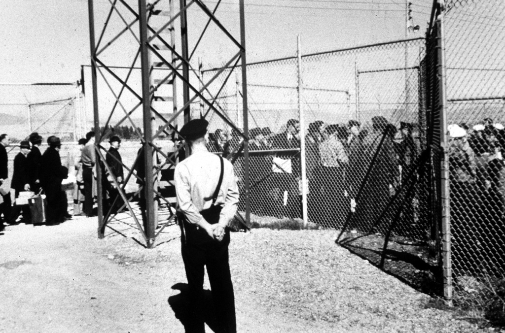 Immigrants arrested as enemy aliens during World War Two passing through a chainlink fence to enter the Fort Missoula internment camp while a sentry stands guard.