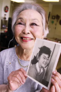 Esther Takei Nishio in 2010 smiling as she holds up a photo of herself taken in the 1940s.