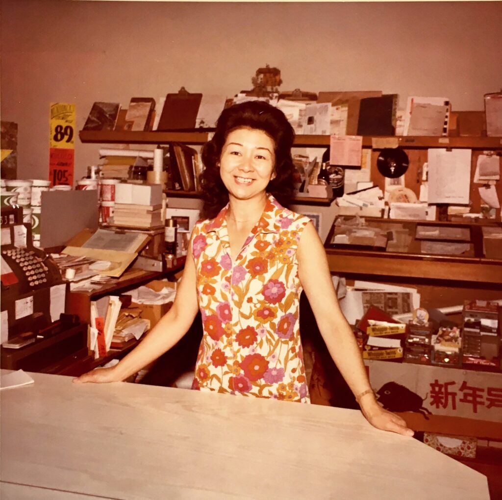 Mary Kageyama Nomura standing behind the counter of her family business, circa 1970s. She is smiling and wearing a brightly colored dress with a floral print.