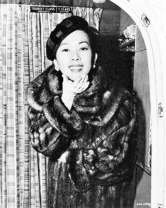 Miiko Taka posing wearing a hat and fur coat. She is smiling and slightly leaning forward with her head cupped in her hand.