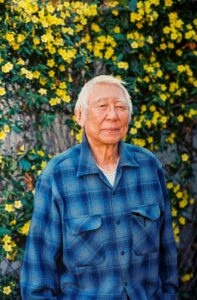 Takashi Hoshizaki standing in front of a wall of yellow flowers with a slight smile.