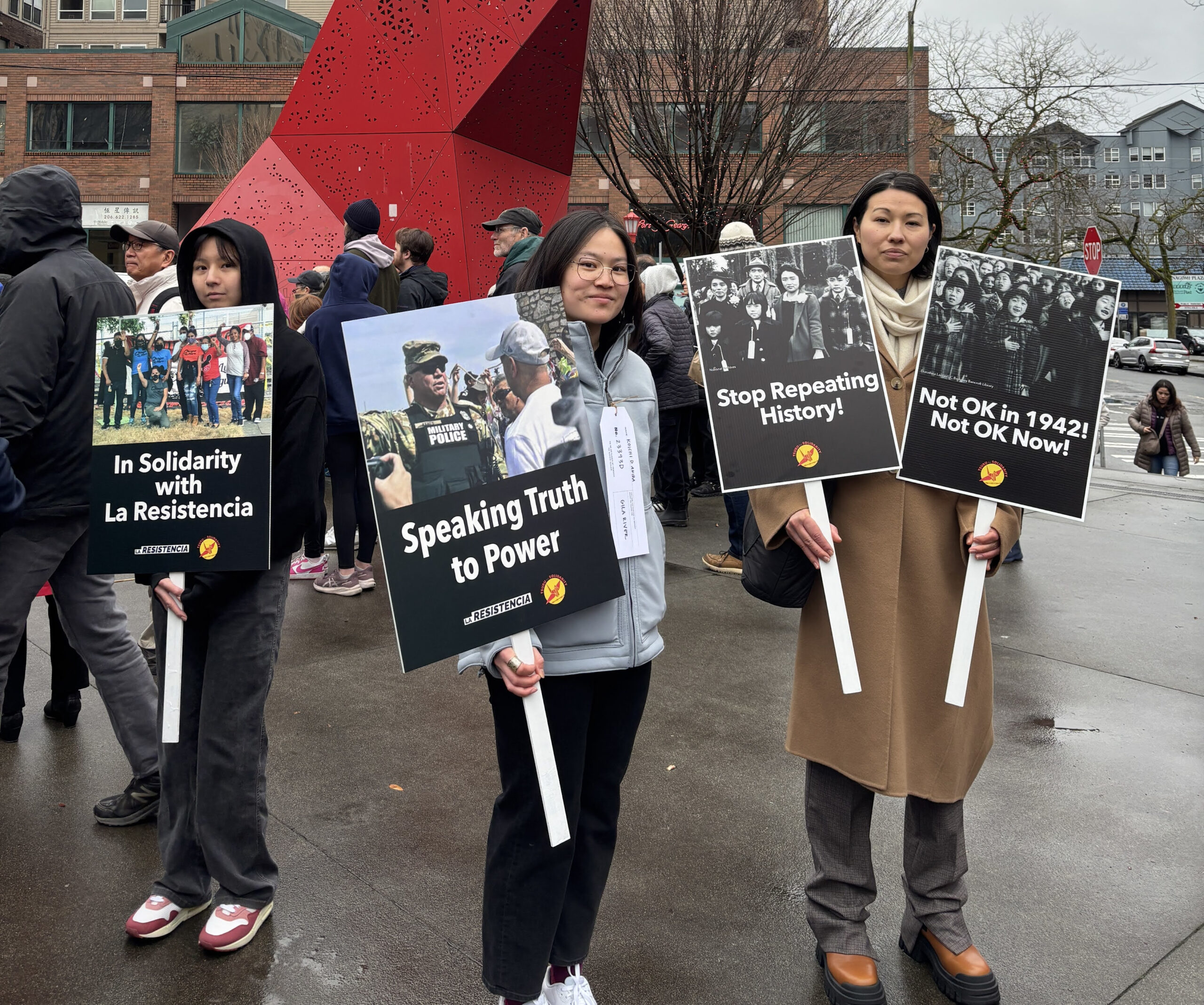 Three Japanese Americans at a Day of Remembrance rally holding up signs with slogans Stop Repeating History, In Solidarity with La Resistencia, Speaking Truth to Power, and Not OK in 1942 Not OK Now.