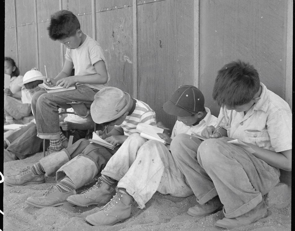 Japanese American students studying in the shade of a barrack at Manzanar concentration camp.
