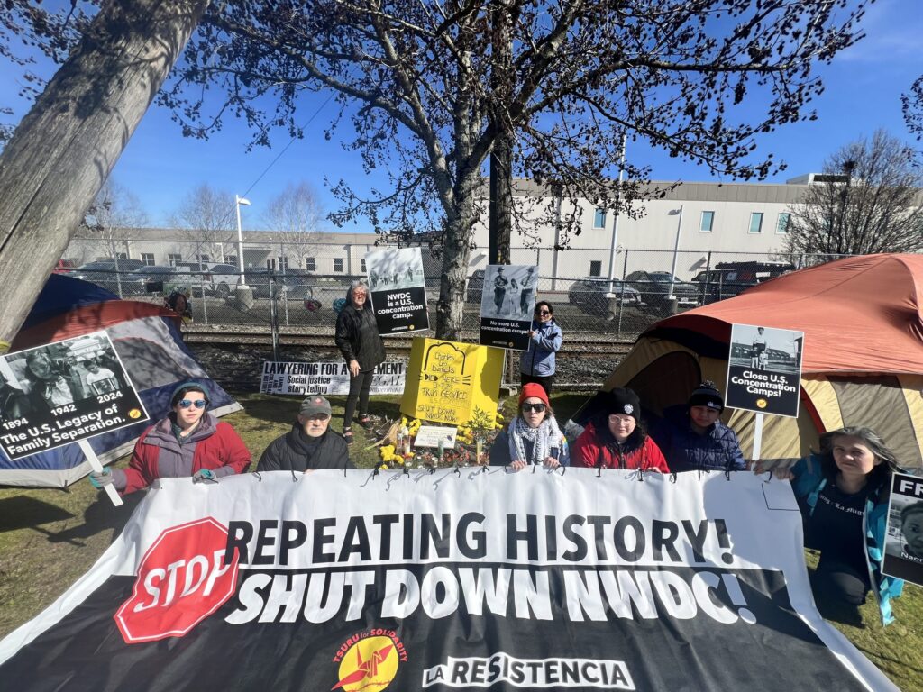 Members of Tsuru for Solidarity stage a protest outside the Northwest Detention Center. Six people in the foreground hold up a banner that reads