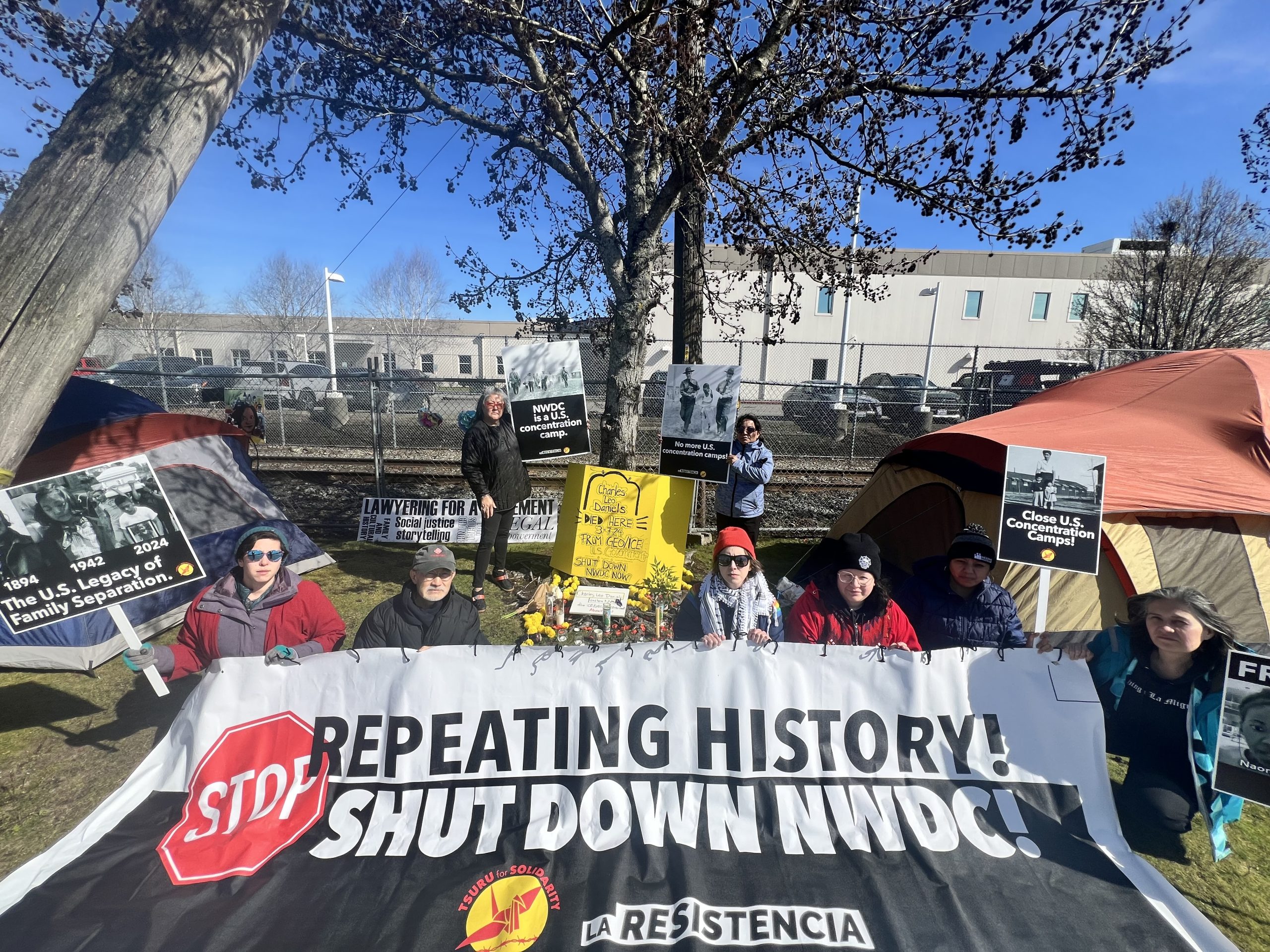 Members of Tsuru for Solidarity stage a protest outside the Northwest Detention Center. Six people in the foreground hold up a banner that reads "Stop Repeating History! Shut down NWDC.!" Two people behind them stand in front of a fence holding signs that say "No more US concentration camps."