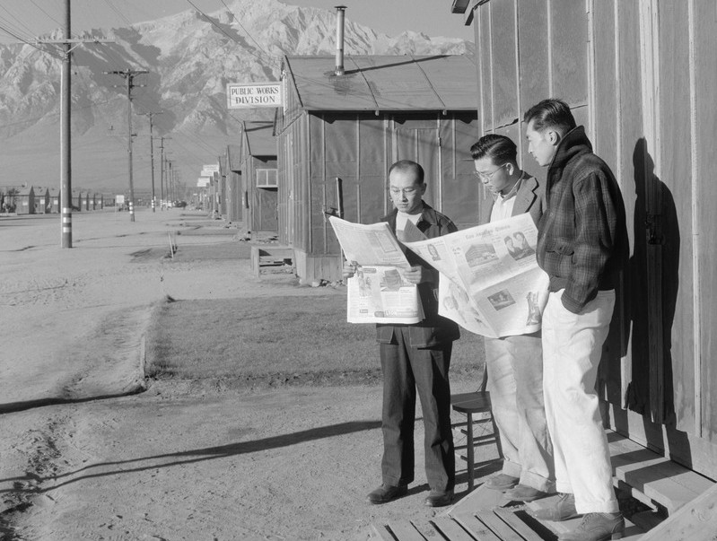 Three Japanese Americans standing outside a barrack in Manzanar reading a newspaper