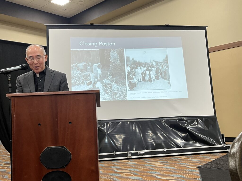 Brian Niiya presenting at the Poston Pilgrimage, standing behind a podium with a presentation slide projected onto a screen behind with the text, "Closing Poston" and two black and white images.