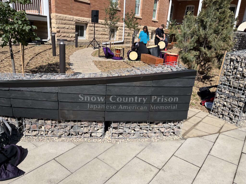 The Snow County Prison Japanese American Internment Memorial wall in North Dakota with a couple of people in the background gathered around drums. 