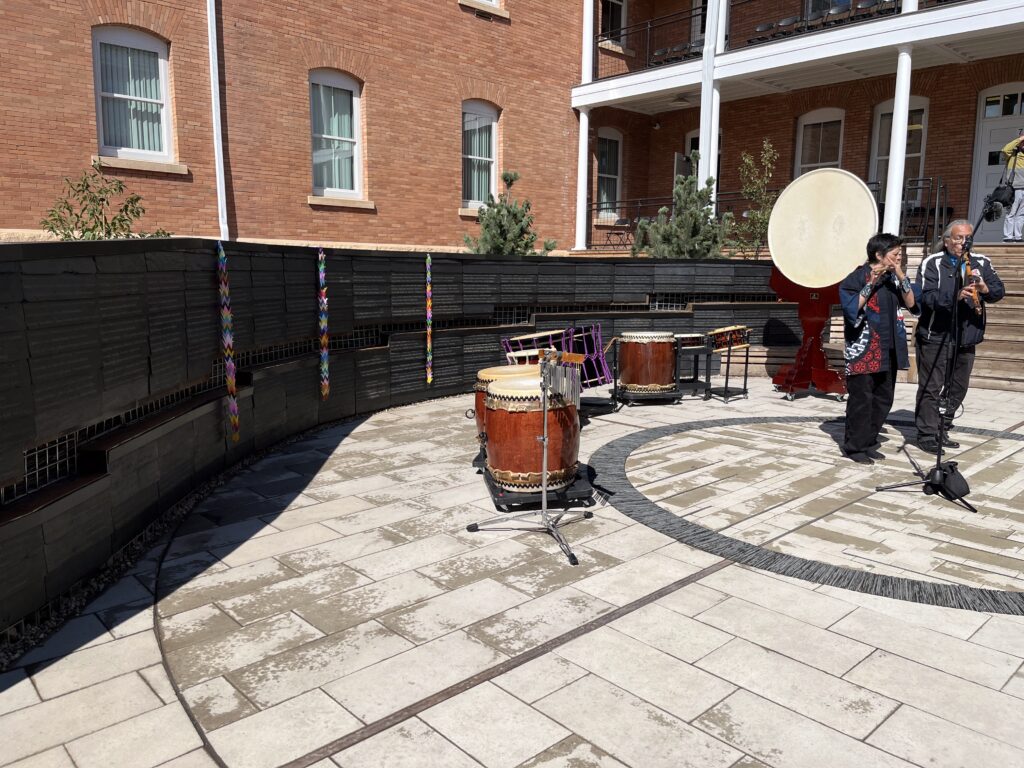 A performance by two people playing instruments. Behind them are taiko drums in a half circle and the Snow County Prison Japanese American Internment Memorial wall.